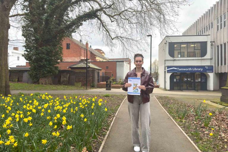 A 16-year-old girl wearing jeans and a leather jacket, standing to the right of a grassy area filled with daffodils, holding a picture of a Staffy outline, which she has coloured in with her winning design.