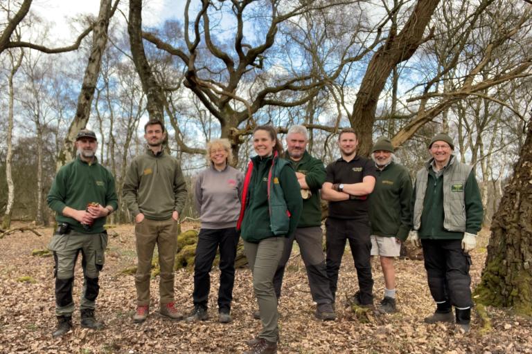 Eight people standing in a woodland area, smiling at the camera, wearing green rangers uniform, gardening gloves and hiking boots.