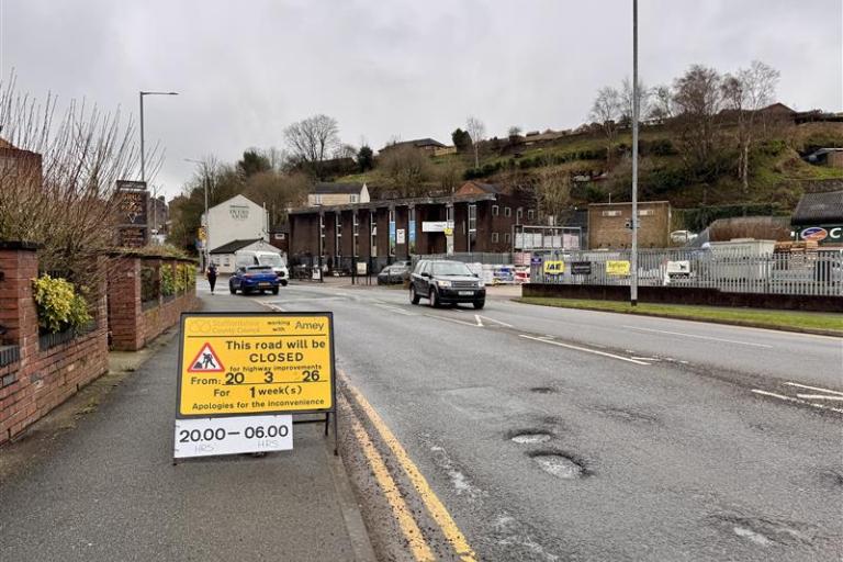 A road with several potholes on it with a temporary yellow sign on the left saying 'this road will be closed' for highways improvements from 20 March for one week between 8pm and 6am.