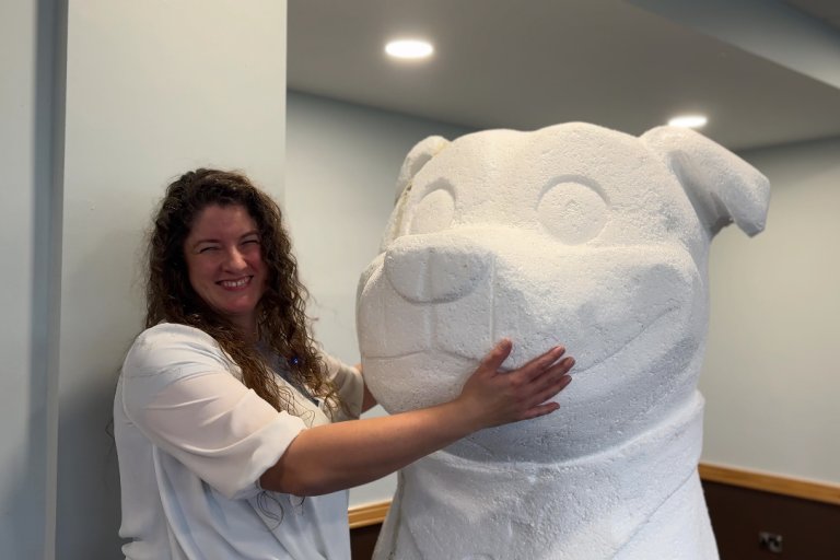 A woman with curly brown hair smiling as she hugs a huge white sculpture of a Staffordshire Bull Terrier dog