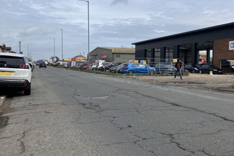 A surface-worn Derby Road in Burton, with cars parked along the left side and businesses on the right side.