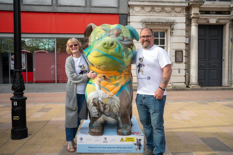 A woman and a man standing either side of a large Staffordshire Bull Terrier sculpture, which has been painted with art inspired by British wildlife and nature.