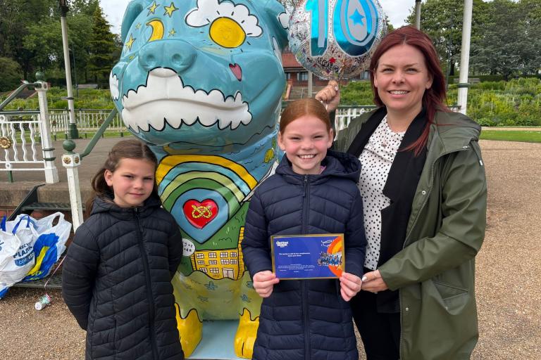 Two young girls standing with their mother next to a Staffordshire Bull Terrier sculpture covered in art including clouds, sunshine, a rainbow and famous Stoke-on-Trent buildings. 