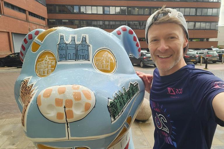 A man dressed in running gear standing with a blue painted Staffordshire Bull Terrier sculpture with artwork on it.