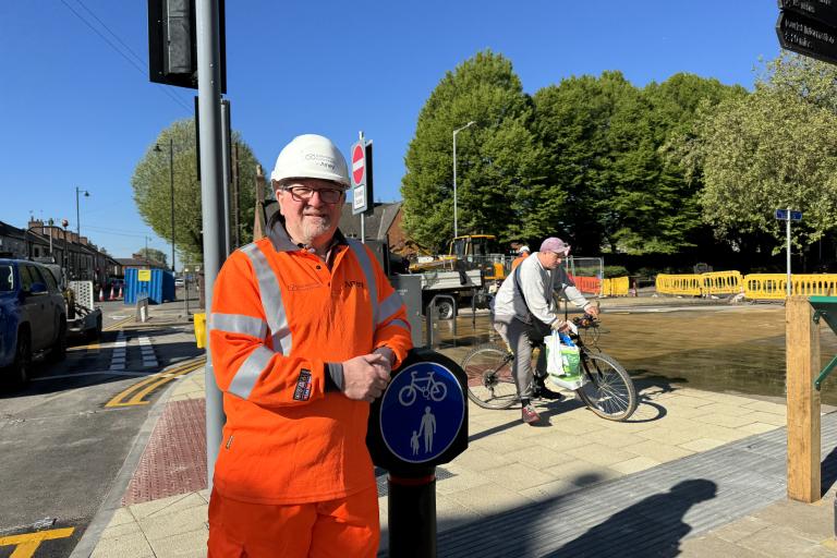 A man in orange high vis gear leaning on a post with a cycleway and footway sign on it. There is also a man on a bike in the background.