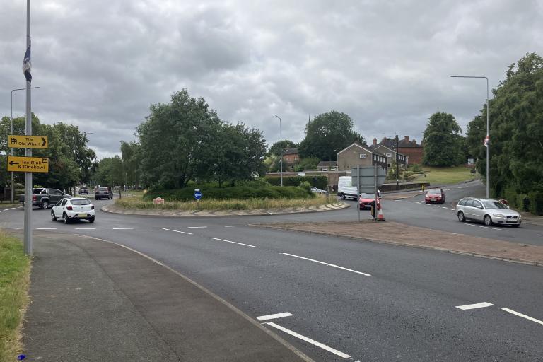 A roundabout with a smooth newly laid road with freshly painted markings and cars driving on the roundabout.