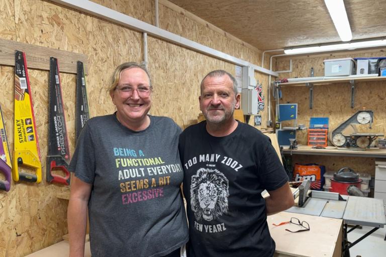 A woman and a man inside a workshop surrounded by tools, including hand saws hanging up on the wall behind them.
