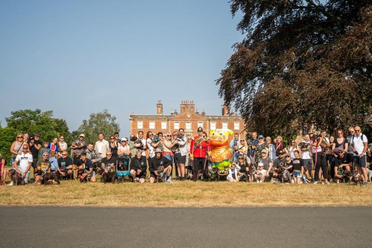 A long line of people with their dogs standing on a big grassy area with a grand looking house in the background on a sunny day.