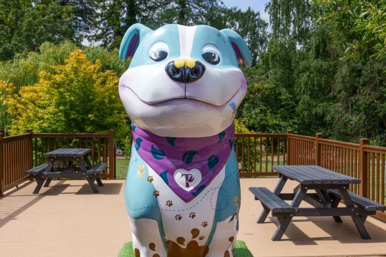 A large turquoise sculpture of a Staffordshire Bull Terrier with colourful artwork painted onto it, including a purple bandana around its neck. It is placed in an outdoor courtyard with trees and picnic benches behind.