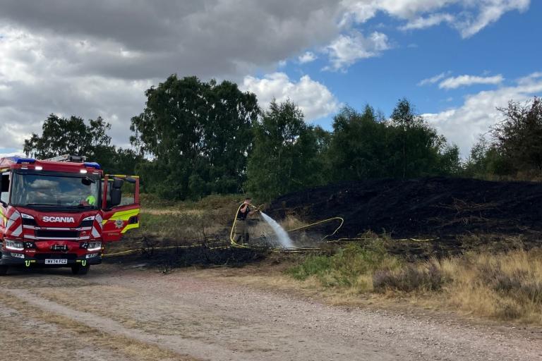A red fire engine pulled up at the side of the road. A firefighter is holding onto a hosepipe, which is connected to the fire engine and hosing down a blackened area of heathland in the countryside. Above, the sky is bright blue with some grey clouds gathering to the left.