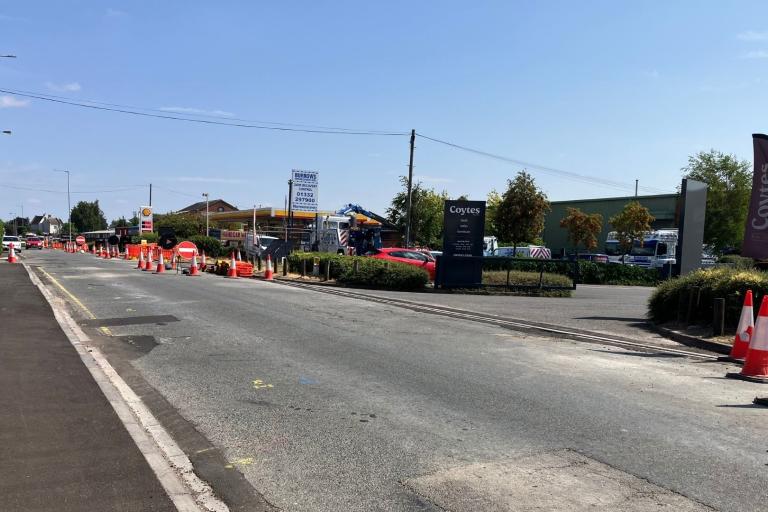 An empty road that has had some patch repairs along the sides, with some orange traffic cones closing off the right lane. To the right of that are some shops.