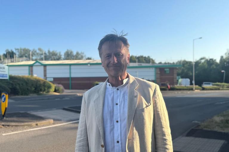 A man standing front and centre, smiling at the camera, whilst stood next to a road.