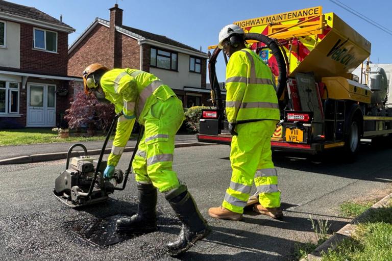 Two highways workers wearing bright yellow jackets and trousers. They're using a wacker machine to flatten down a pothole they have just filled. A big yellowy-orange highways truck can be seen in the background. It is a very hot and sunny day.
