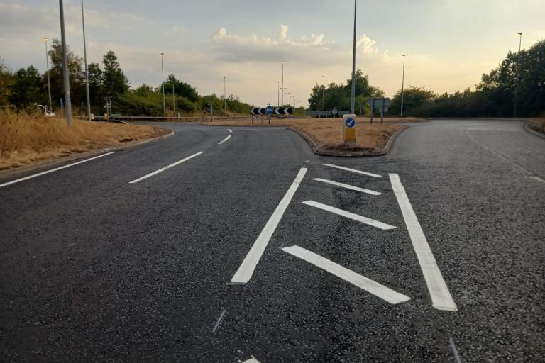 Image shows a lovely, flat, newly resurfaced  road leading up to a roundabout. There are hatch markings and white lines visible. The image is taken late evening so the sun is low and basking everything in an ethereal glow.