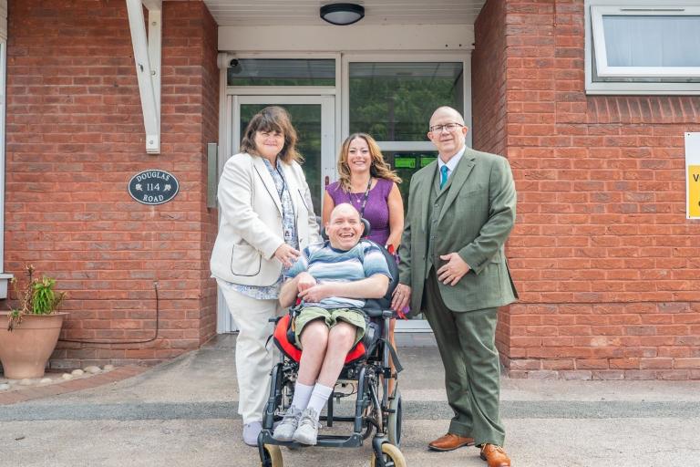 Two smartly dressed women and a man standing behind a man in a wheelchair. They are all smiling as they are stood outside the entrance to a brick building.