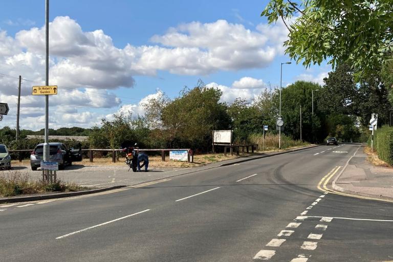A four-way junction in Burton, with a bus stop and parking area to the right of the road. It is a sunny day.