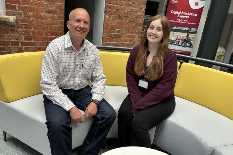 A smartly dressed smiling man and woman sitting down on a modern curved yellow and grey sofa.