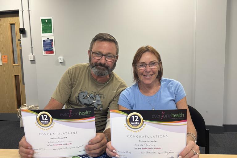 Andrew and Michelle sitting at a table holding certificates from Everyone Health recognising them for being smoke-free for 12 weeks. The certificates are displayed towards the camera in an indoor meeting room setting.