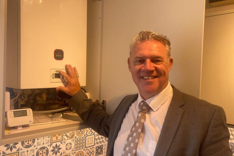 Anthony Screen stands beside a domestic boiler, pointing to the controls.