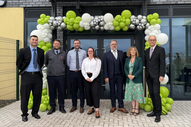Group of seven people standing outside Greywood school entrance beneath a green, white and silver balloon arch during a school opening or celebration event.