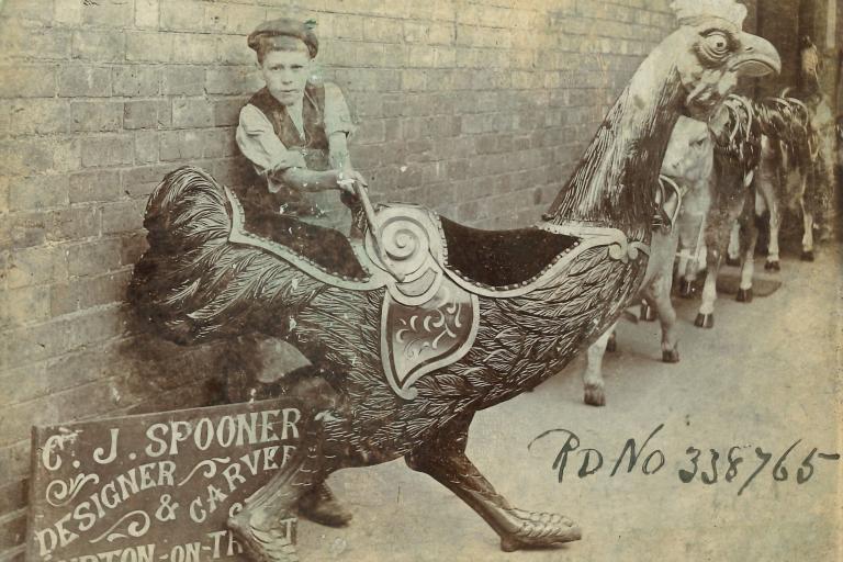 Historical photograph showing a young boy posing beside a carved fairground cockerel manufactured by C.J. Spooner.