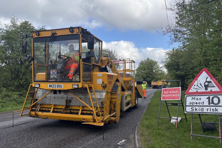 Road resurfacing works on a rural road, with a yellow paving machine, road workers in high-visibility clothing, and warning signs advising of no road markings and a 10mph maximum speed.