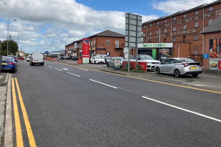 A smooth newly resurfaced road with freshly painted double yellow lines running along the side of it and several vehicles driving along the route. It is a bright day with blue sky peeking through the clouds in the sky.