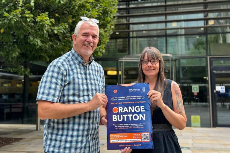 A smiling man in a checkered t-shirt standing with a woman with dark hair and a dark dress in front of a building outside and holding a poster promoting the 'Orange Button' scheme.