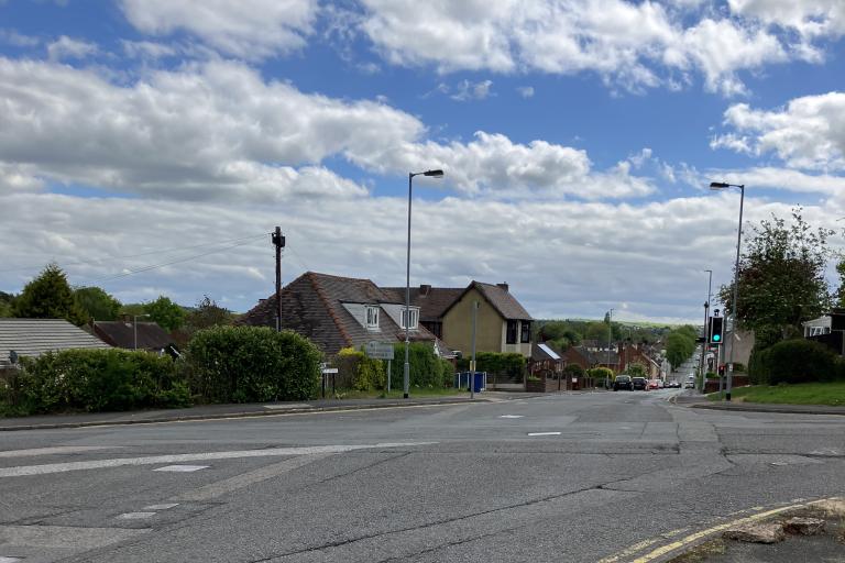 A picture of a crossroads with houses in the middle distance on a cloudy day. The road looks tired and in need of refurbishment.