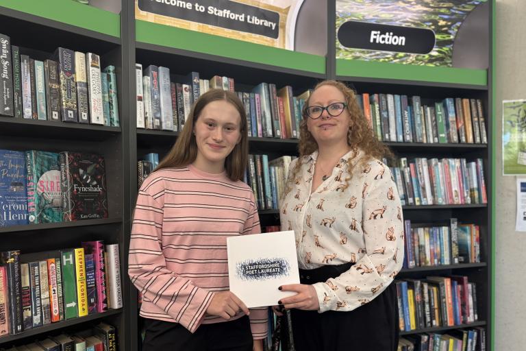Lola (left) and Cherry (right) stand in front of bookshelves in Stafford Library, holding a Staffordshire Poet Laureate folder beneath the library's fiction section signage.
