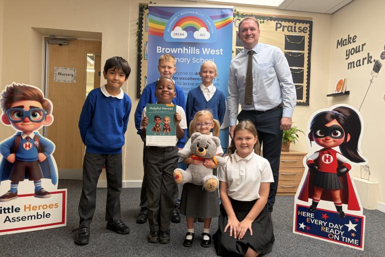 Pupils and staff at Brownhills West Primary School pose with a Little Heroes resource booklet and teddy bear, standing between two superhero character displays in a school classroom.