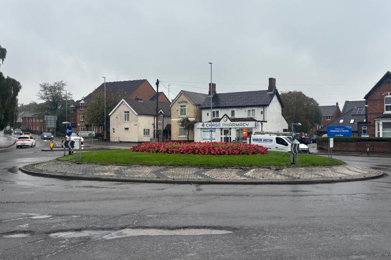 A picture of a roundabout on a grey, rainy day. The roundabout has red flowers on it. There is a white house in the background.