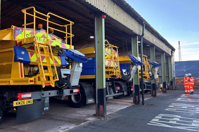 A row of three gritting machines in a depot shed. You can see the rear of the gritters, and the spinner where the salt comes out. There are two men in bright orange in the far right of the photo.