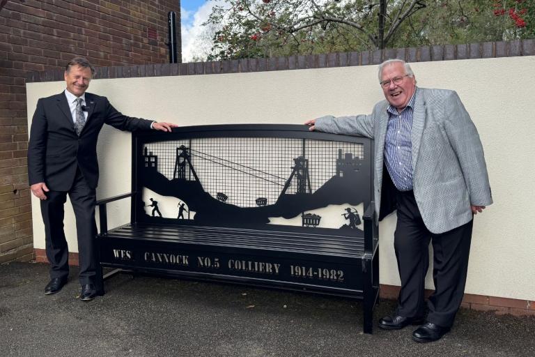 A black steel bench with mining designs on the backrest. There are two men, smartly dressed in suits, smiling and leaning on each side of it.