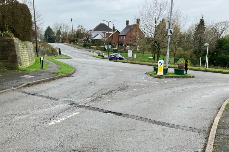 A surface-worn road approaching a roundabout. The day is overcast.