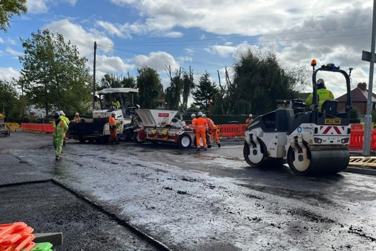 A group of highways workers, wearing bright orange and yellow hi-vis and white hard hats, working on repairing a road with various bits of machinery, including a roller. The work site is surrounded by bright orange plastic fencing and there is a bright blue sky above them with some clouds.