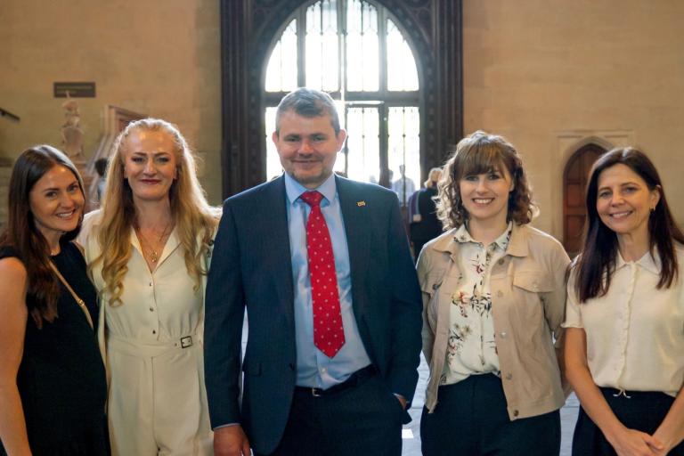 Four smartly dressed women and a man standing together and smiling at the camera. They are indoors and, behind them, is a staircase and an arched window that stretches from the ceiling to the floor.
