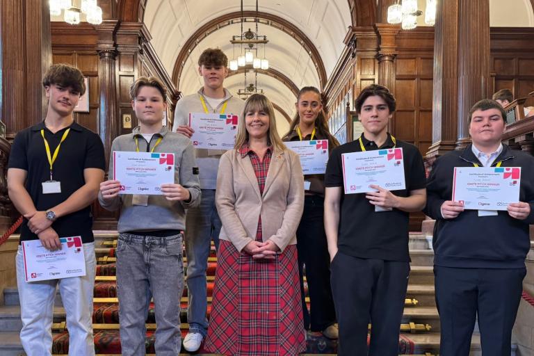 Five students facing forward, smiling, and holding certificates. A smiling woman in a red tartan dress and brown jacket is standing in the centre of them. They are indoors, standing on a wide red carpeted staircase. 