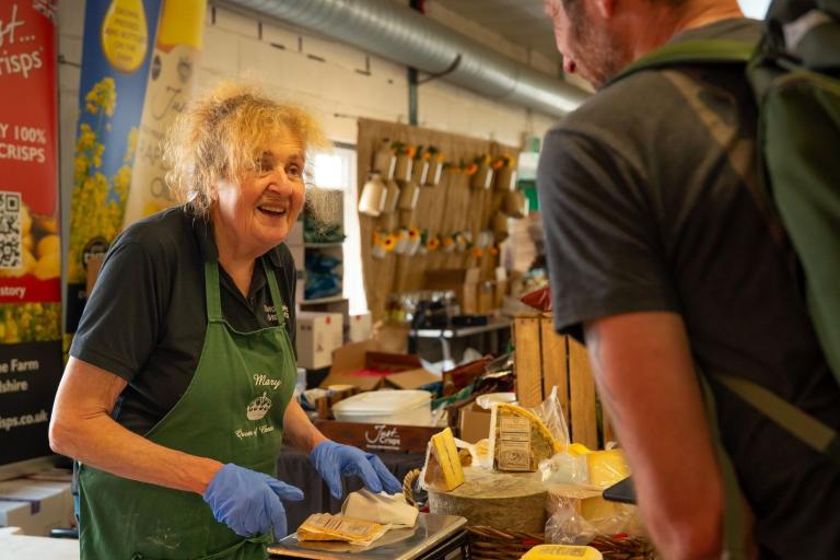 A smiling woman, wearing a green apron and blue gloves serving cheese to a male customer.