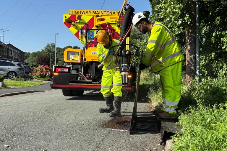 Two highways workers in bright yellow, next to a large highways machine, repairing a road.