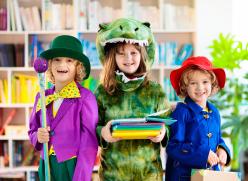 Three children in colourful costumes standing in a library, with one holding a stack of books.