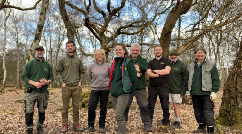 Eight people standing in a woodland area, smiling at the camera, wearing green rangers uniform, gardening gloves and hiking boots.