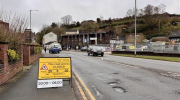 A road with several potholes on it with a temporary yellow sign on the left saying 'this road will be closed' for highways improvements from 20 March for one week between 8pm and 6am.