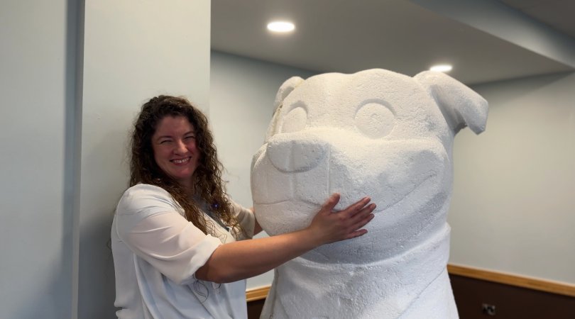 A woman with curly brown hair smiling as she hugs a huge white sculpture of a Staffordshire Bull Terrier dog