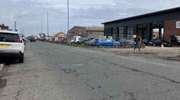 A surface-worn Derby Road in Burton, with cars parked along the left side and businesses on the right side.