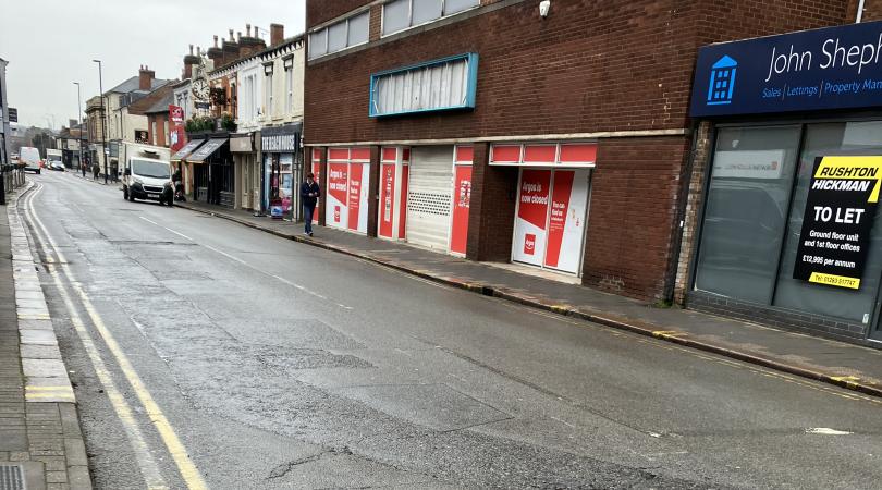 A surface-worn road in Burton town centre, with businesses running along the right side of the road.