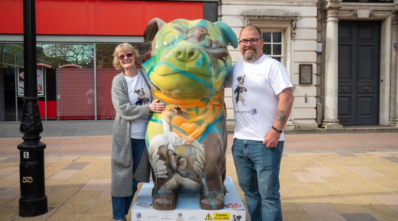 A woman and a man standing either side of a large Staffordshire Bull Terrier sculpture, which has been painted with art inspired by British wildlife and nature.