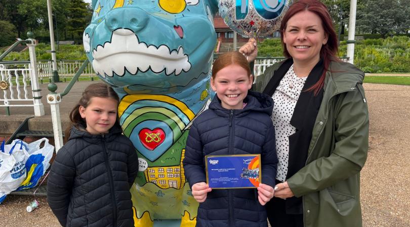 Two young girls standing with their mother next to a Staffordshire Bull Terrier sculpture covered in art including clouds, sunshine, a rainbow and famous Stoke-on-Trent buildings. 
