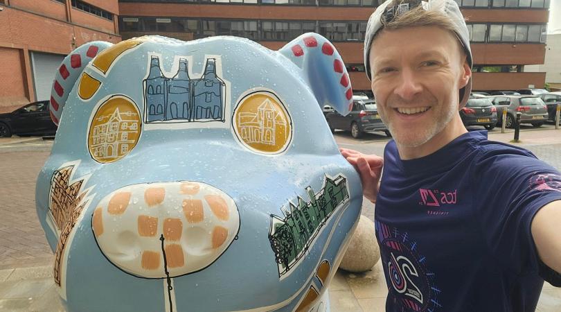 A man dressed in running gear standing with a blue painted Staffordshire Bull Terrier sculpture with artwork on it.