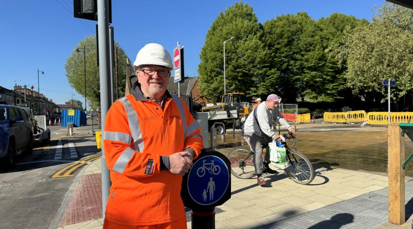 A man in orange high vis gear leaning on a post with a cycleway and footway sign on it. There is also a man on a bike in the background.
